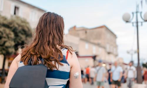 Back view of woman outdoors with a CGM on her upper arm & wearing a backback
